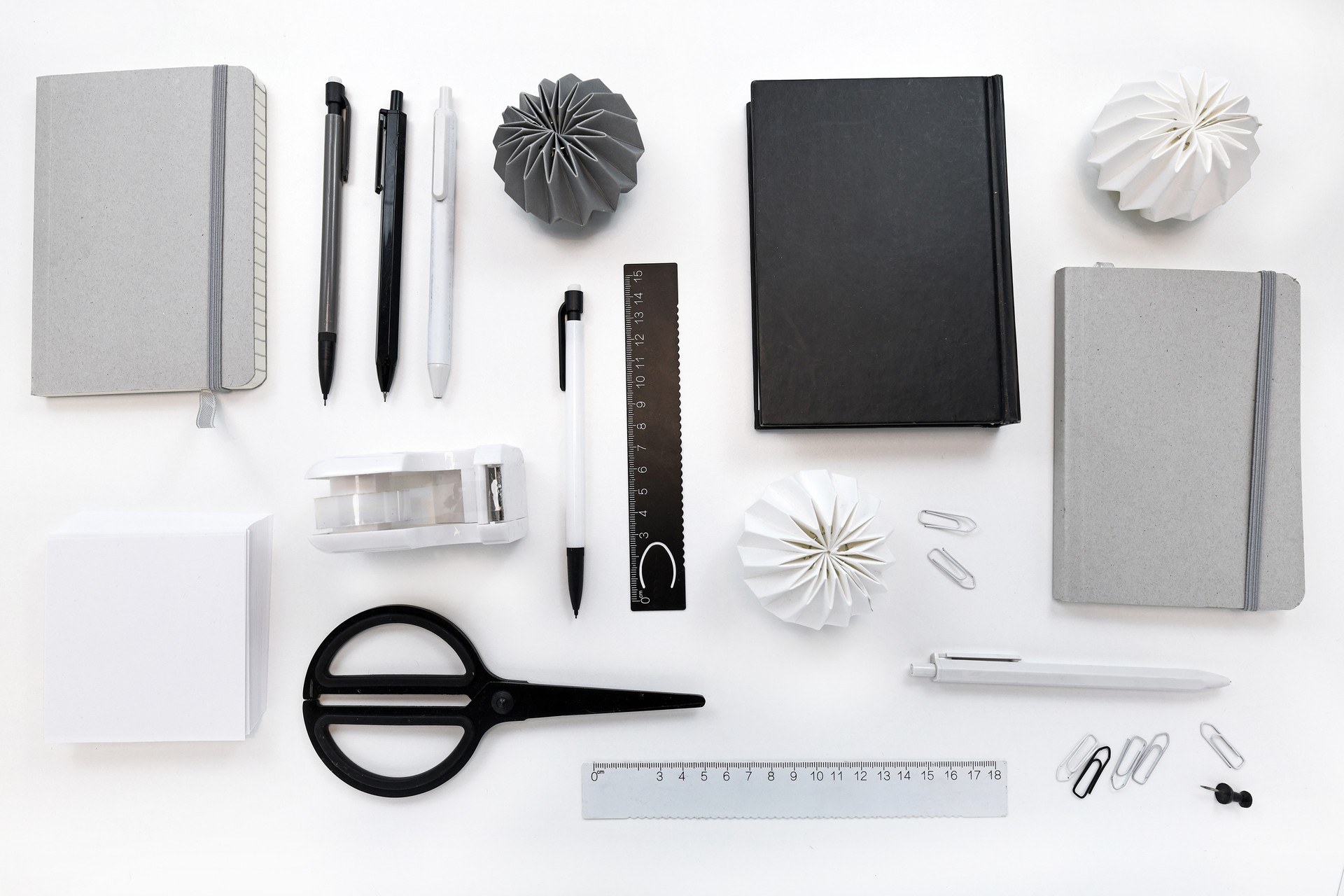 High angle top view of white desk with various black, gray and white neatly arranged office supplies like scissors, pens, pencils and notebooks